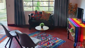 A living room with a sofa, two chairs, a small round table, a colorful rug, a TV stand, and a window with curtains. - El Aguacatal (Panajachel)