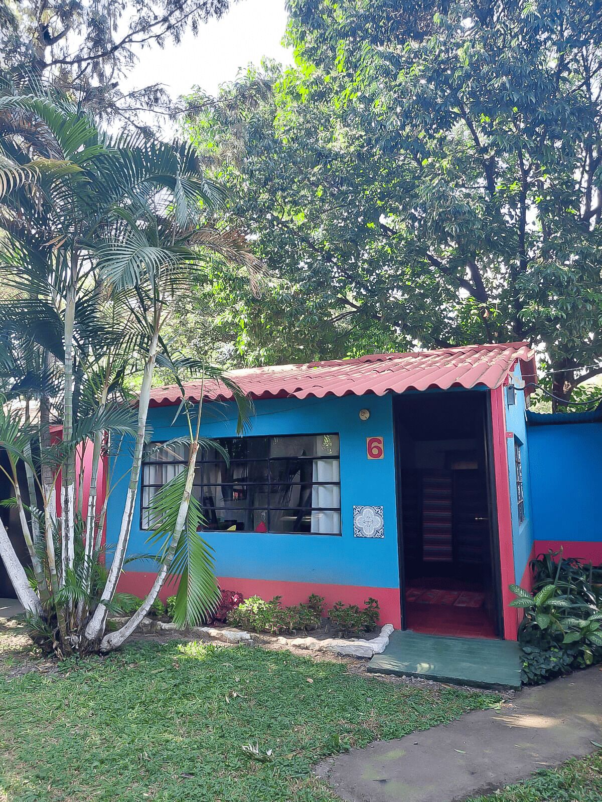 A small, single-story building with a red roof and blue walls, surrounded by lush greenery.