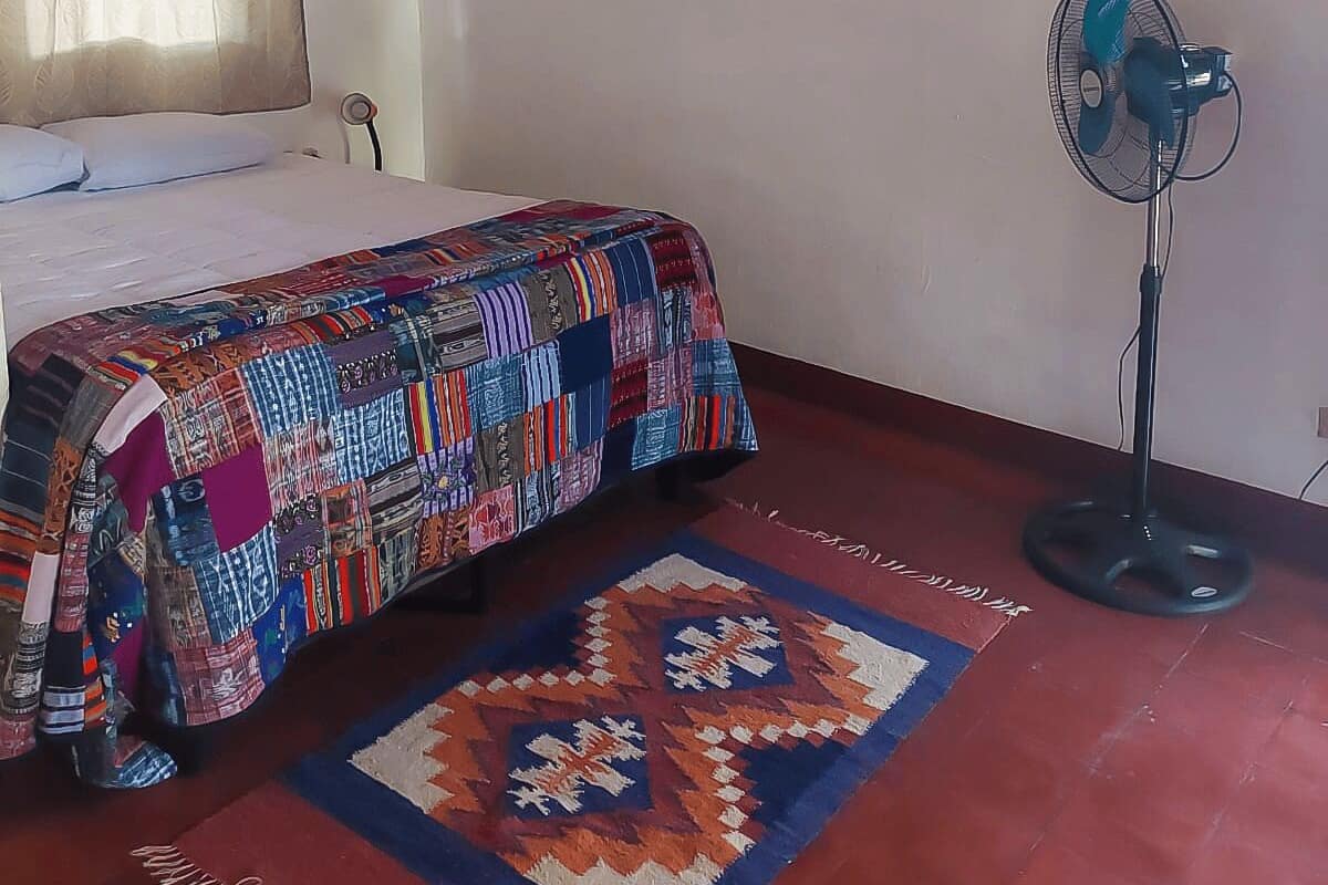 Une chambre à coucher avec un lit, une couette, un tapis, un tableau accroché au mur et un ventilateur sur pied.