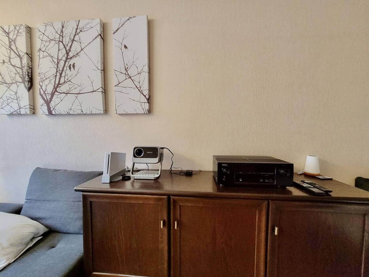 A living room with a wooden cabinet, a projector, a speaker, and a remote control on a table. There is a grey sofa with a pillow and a wall-mounted picture frame.