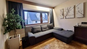Un salon moderne avec un canapé d’angle, une plante en pot, une grande fenêtre avec des rideaux et des œuvres d’art accrochées au mur.