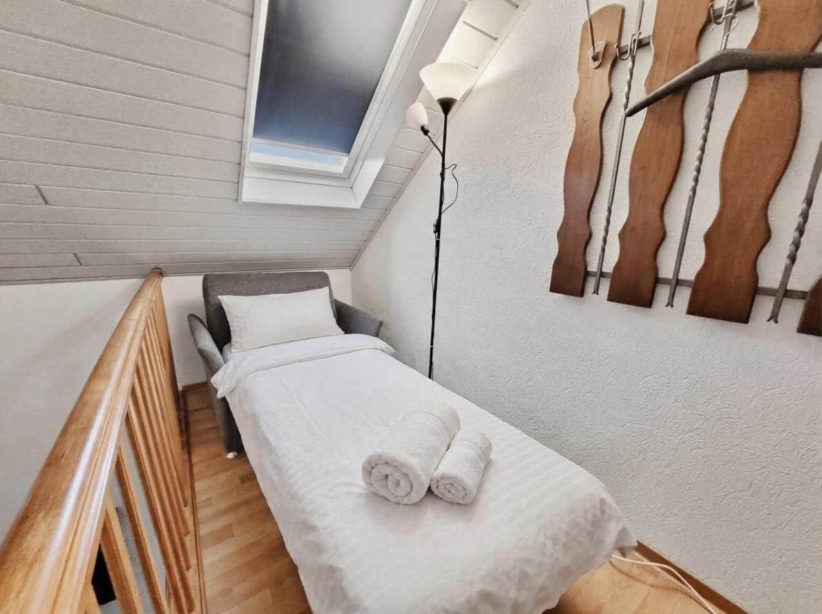 A small, single-bed room with a skylight, a wooden headboard, and a wall-mounted light fixture.