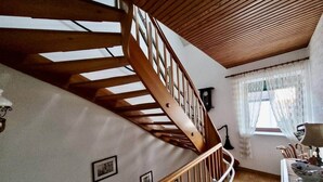 Un escalier en bois, doté d’une rampe, mène à une pièce où se trouvent une fenêtre, une lampe et une table.