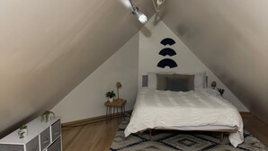 Ett litet, ombonat sovrum på vinden med ett sluttande tak, en säng med ett vitt täcke, en mönstrad matta och ett trägolv.