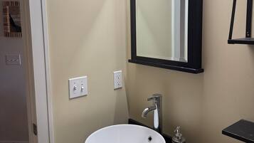 Een badkamer met een witte wastafel, een zwart aanrecht en een spiegel met een zwarte lijst. Er zijn twee lichtschakelaars aan de muur.