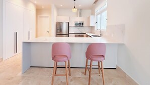 A modern kitchen with a central island, stainless steel appliances, and two pink bar stools. - Tropical Voyage 3BR Garden View (Rio Hato, Coclé)