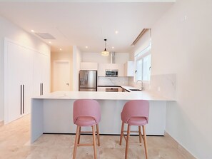 A modern kitchen with a central island, stainless steel appliances, and two pink bar stools. - Tropical Voyage 3BR Garden View (Rio Hato, Coclé)