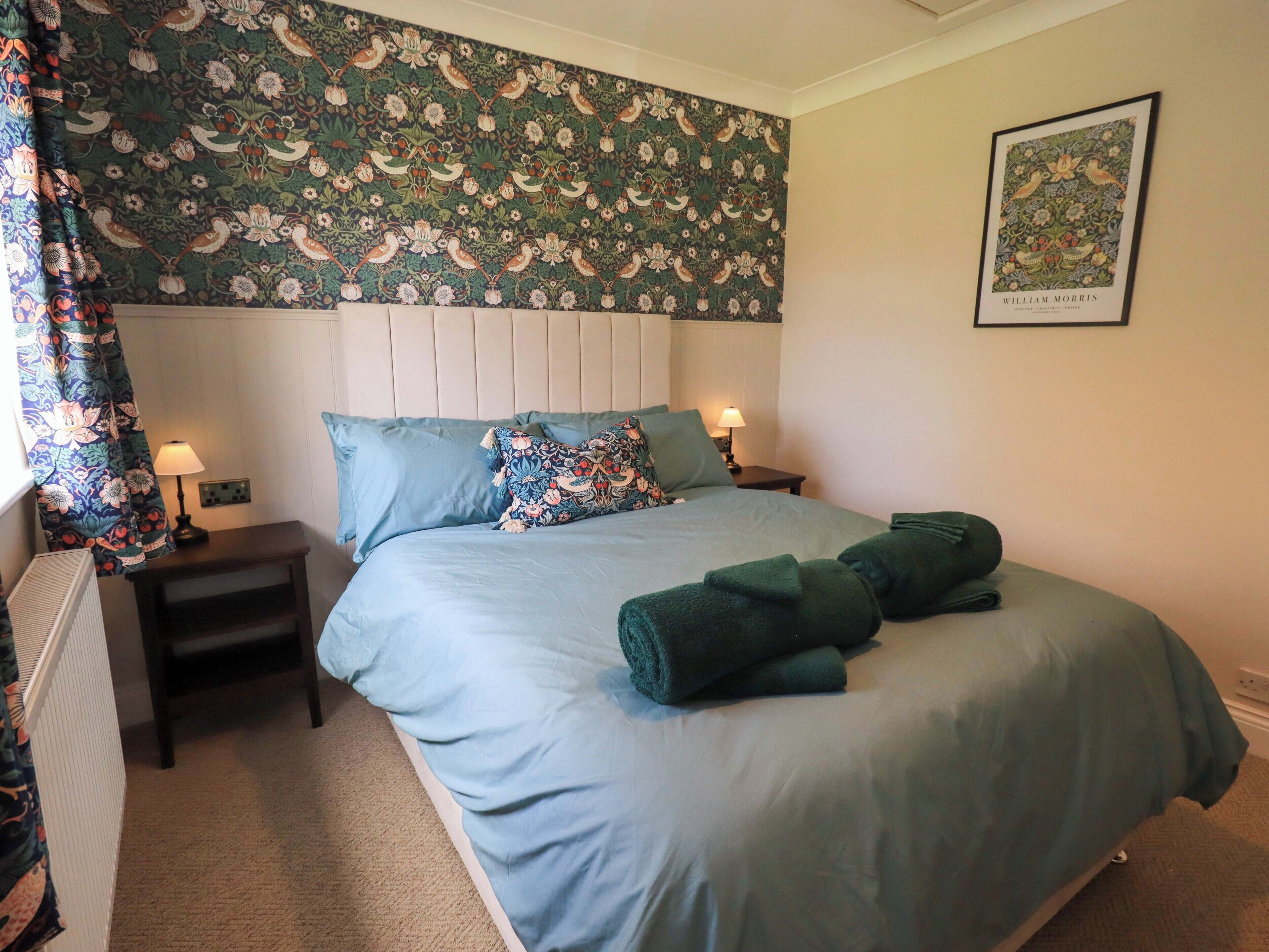 A bedroom with a floral wallpaper, a bed with blue bedding, two bedside tables with lamps, and a framed picture on the wall.