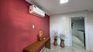 Een gang met een rode muur, een houten bankje, twee terracotta potten met takken, en een airconditioning die aan de muur is bevestigd.