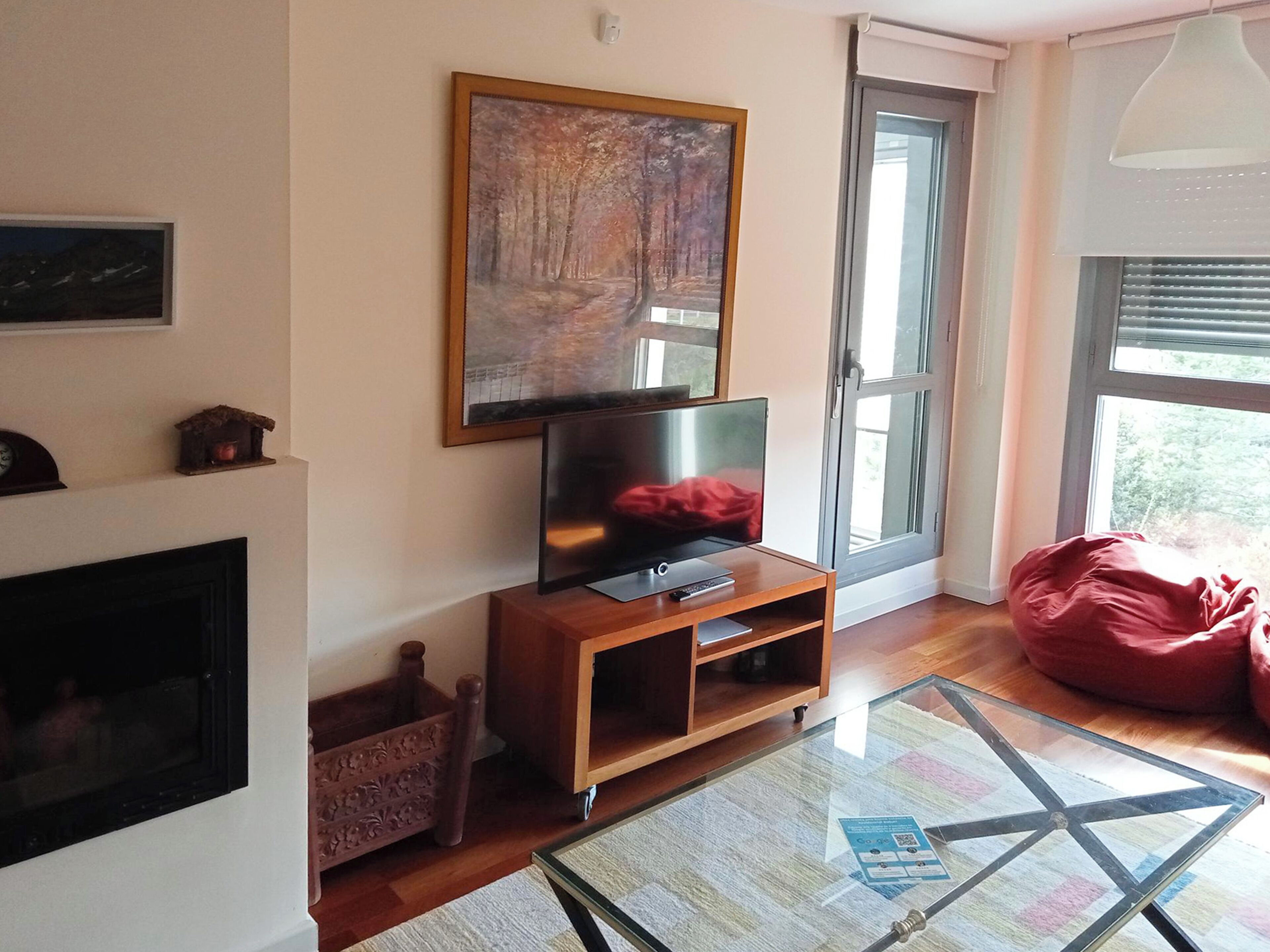 Een woonkamer met een open haard, een televisie op een houten standaard, een schilderij boven de televisie en een glazen salontafel.
