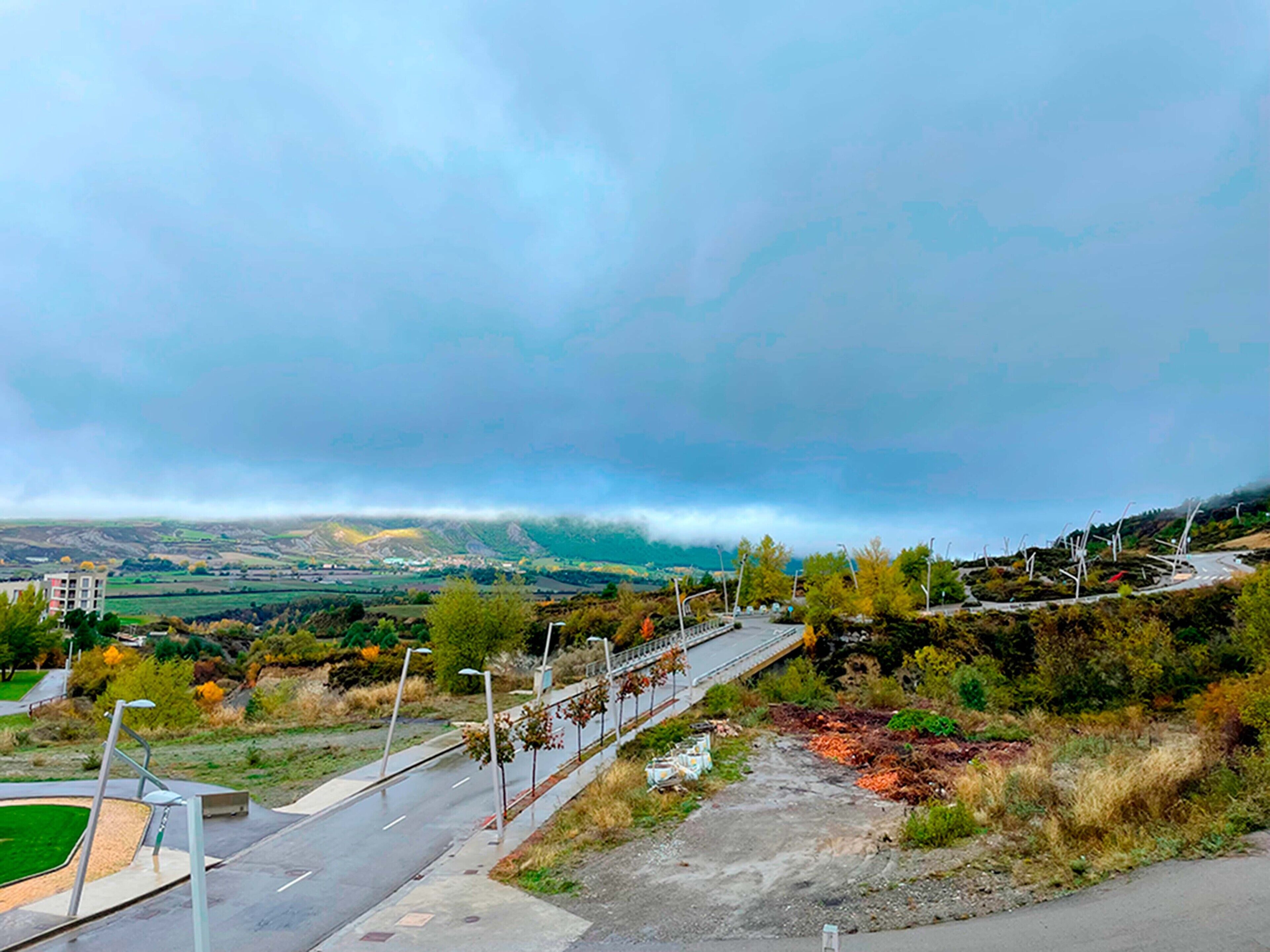 A road with a guardrail, a pedestrian path, and a view of a valley with buildings and trees under a cloudy sky.