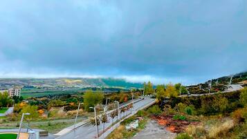 A road with a guardrail, a pedestrian path, and a view of a valley with buildings and trees under a cloudy sky.