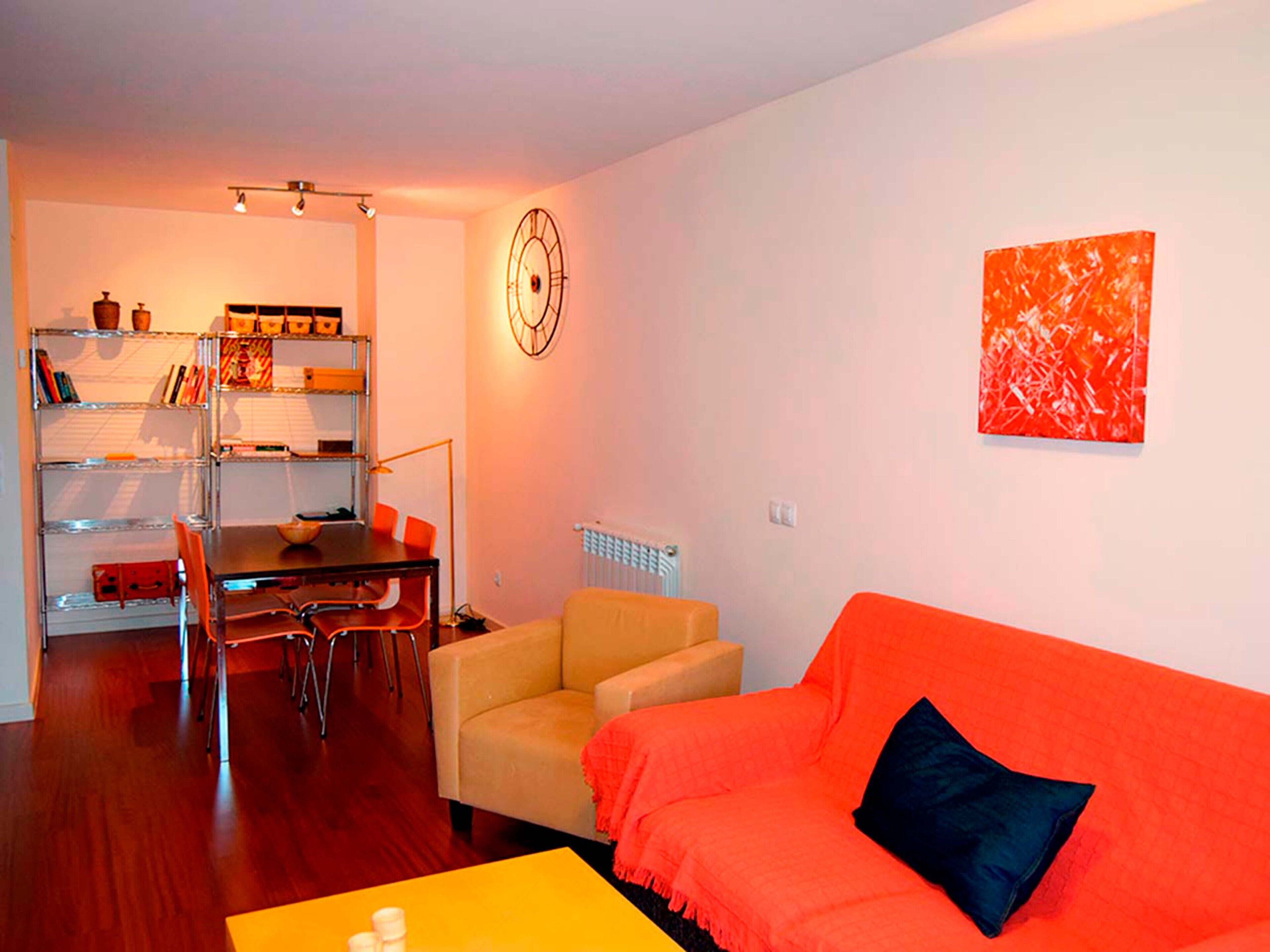 A living room with a wooden floor, a yellow table, an orange sofa with a black pillow, a beige armchair, a dining table with chairs, a bookshelf, and a wall clock.