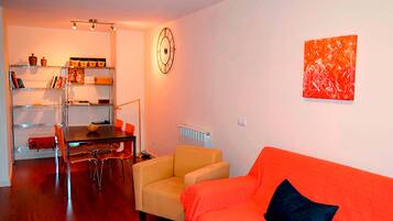 A living room with a wooden floor, a yellow table, an orange sofa with a black pillow, a beige armchair, a dining table with chairs, a bookshelf, and a wall clock.