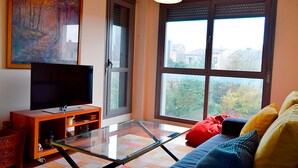 A living room with a glass-top coffee table, a flat-screen TV on a wooden stand, a sofa with colorful cushions, and a large window with a view of trees and houses. - Apartamentos Las Margas Sabiñanigo 3000 (Sabiñánigo)