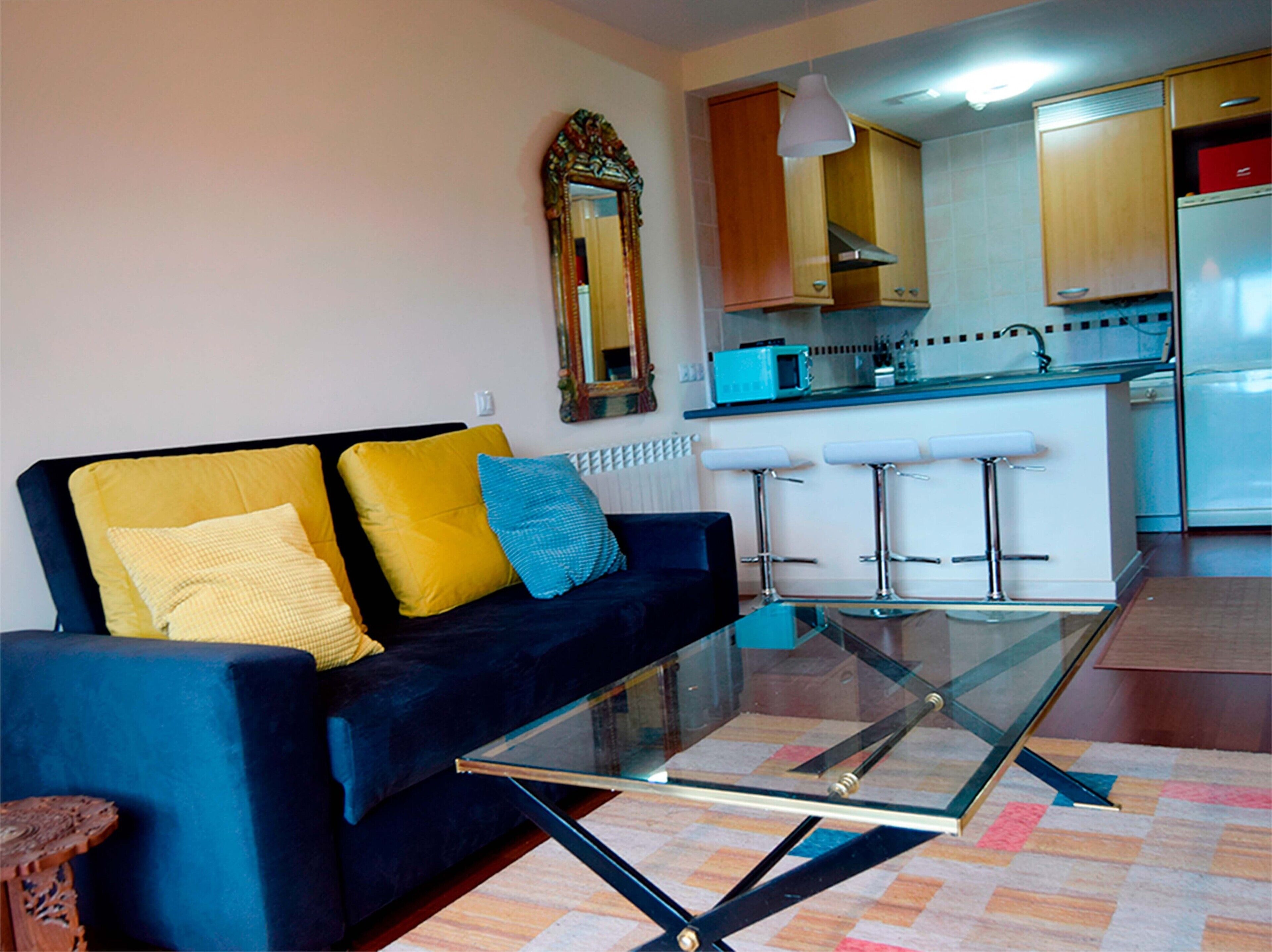 A living room with a dark blue sofa, yellow and blue cushions, a glass coffee table, and a kitchen area with wooden cabinets and a microwave.