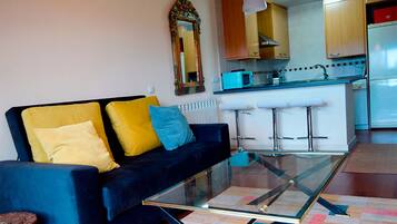 A living room with a dark blue sofa, yellow and blue cushions, a glass coffee table, and a kitchen area with wooden cabinets and a microwave.