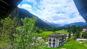 A view of a building with multiple balconies under a bridge, surrounded by green fields and mountains. - Rifugio TEA Dolomiti (Canale d'Agordo)