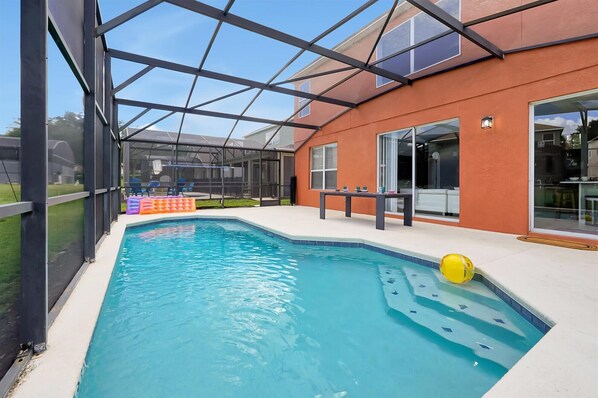 Une piscine entourée de parois en verre, un espace patio aménagé avec du mobilier d’extérieur et un bâtiment doté de grandes fenêtres.