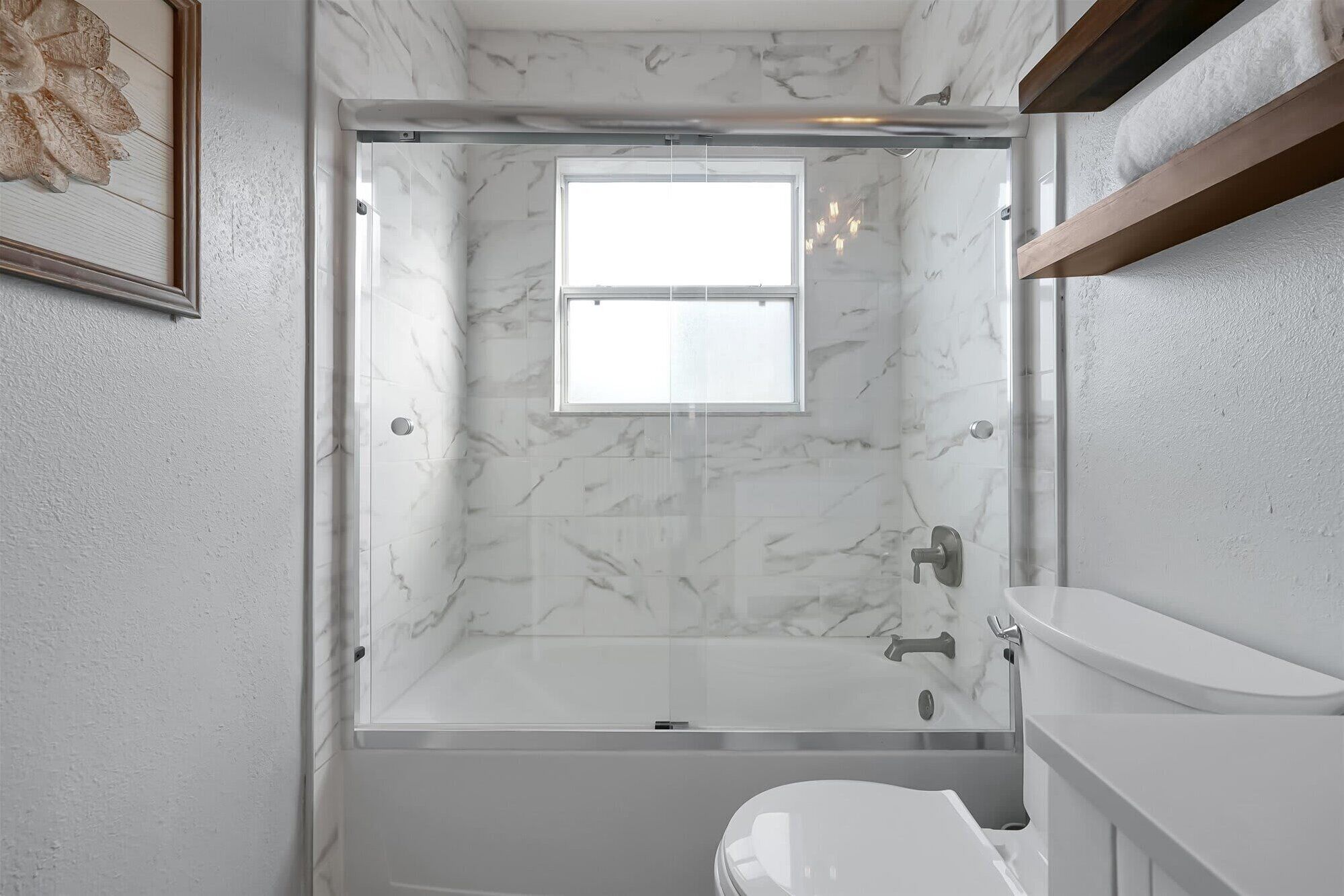 Un baño moderno con ducha envuelta en vidrio, pared de mármol blanco y una ventana pequeña.