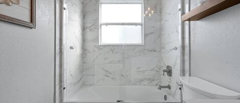 Un baño moderno con ducha envuelta en vidrio, pared de mármol blanco y una ventana pequeña.