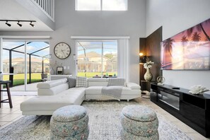 Un salon moderne avec un canapé d’angle, des poufs, une grande horloge et un tableau décoratif représentant un téléviseur.