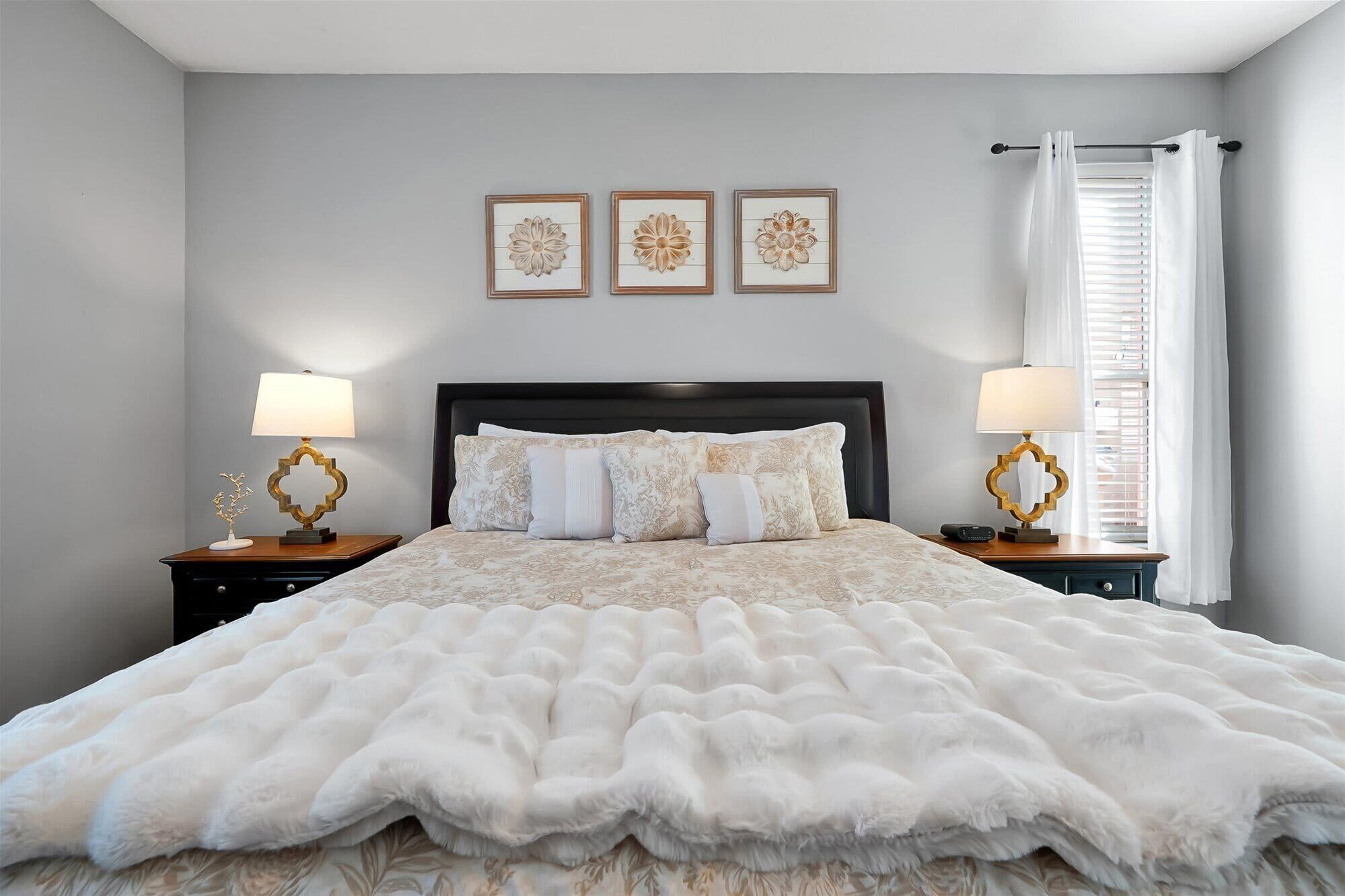 Una camera da letto con un letto grande, due comodini con lampade, una finestra con tende e tre decorazioni floreali a parete.