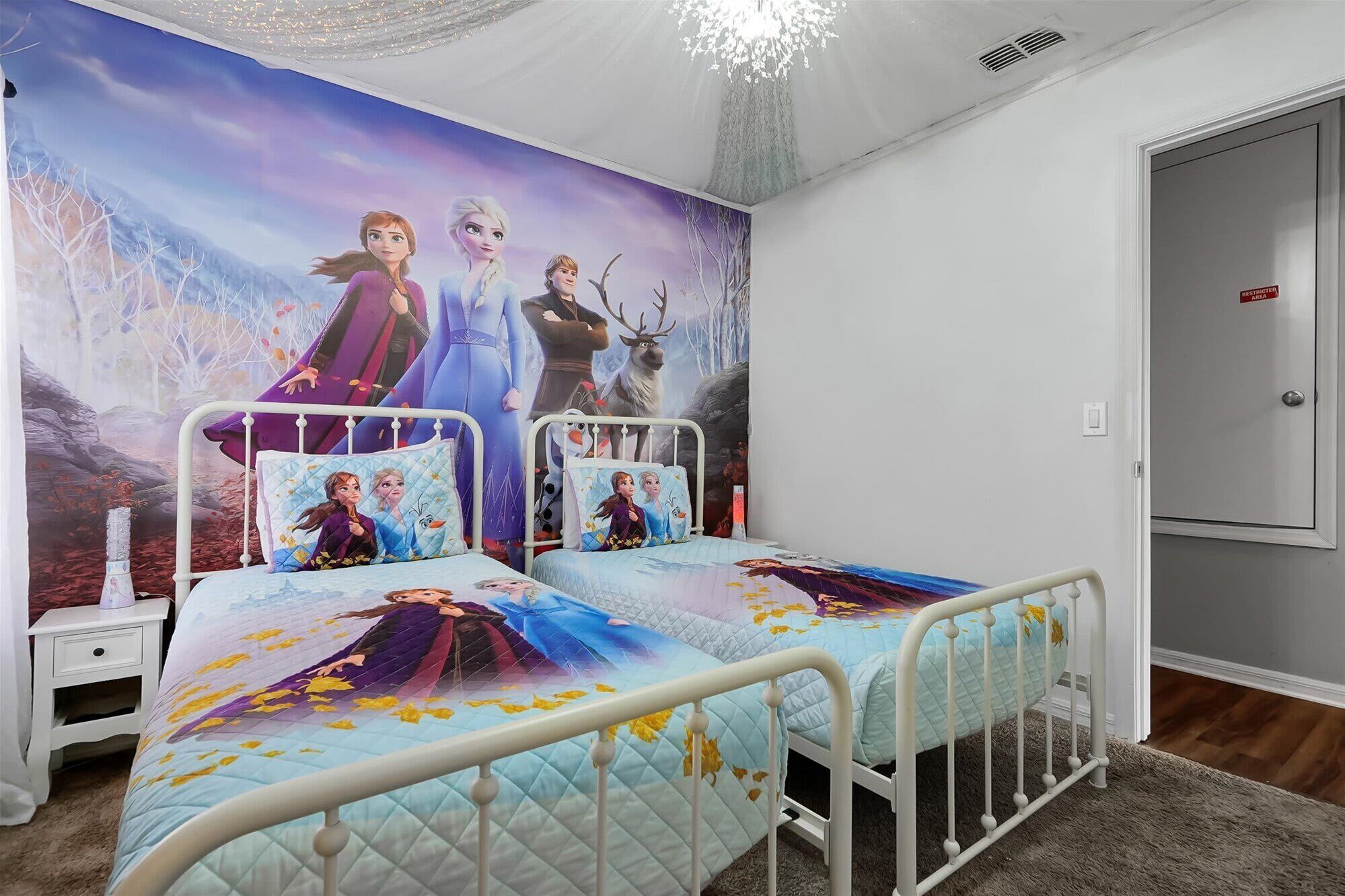 A bedroom with two beds, each with a Frozen-themed bedspread and pillows. There is a nightstand with a lamp and a small table beside one bed. The room has white walls and a large window.