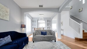 Amplia sala de estar con un sofá azul, un sillón gris y un otoman redonda. Se ve una escalera a la derecha y un comedor al fondo.