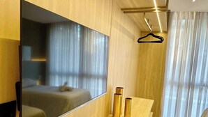 Ein modernes Hotelzimmer mit einem großen Spiegel, einem Bett mit weißer Tagesdecke, einem Holzschminktisch mit zwei goldfarbenen Flaschen und transparenten Vorhängen.