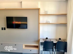 Una habitación moderna con televisor de pantalla plana mostrando 'NETFLIX', un comedor con mesa y sillas, y una pequeña estantería con objetos decorativos.