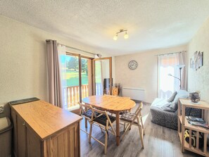 Ein Zimmer mit Esstisch, Stühlen, einer Couch, einem Heizkörper, einer Uhr und einem Fenster mit Vorhängen.