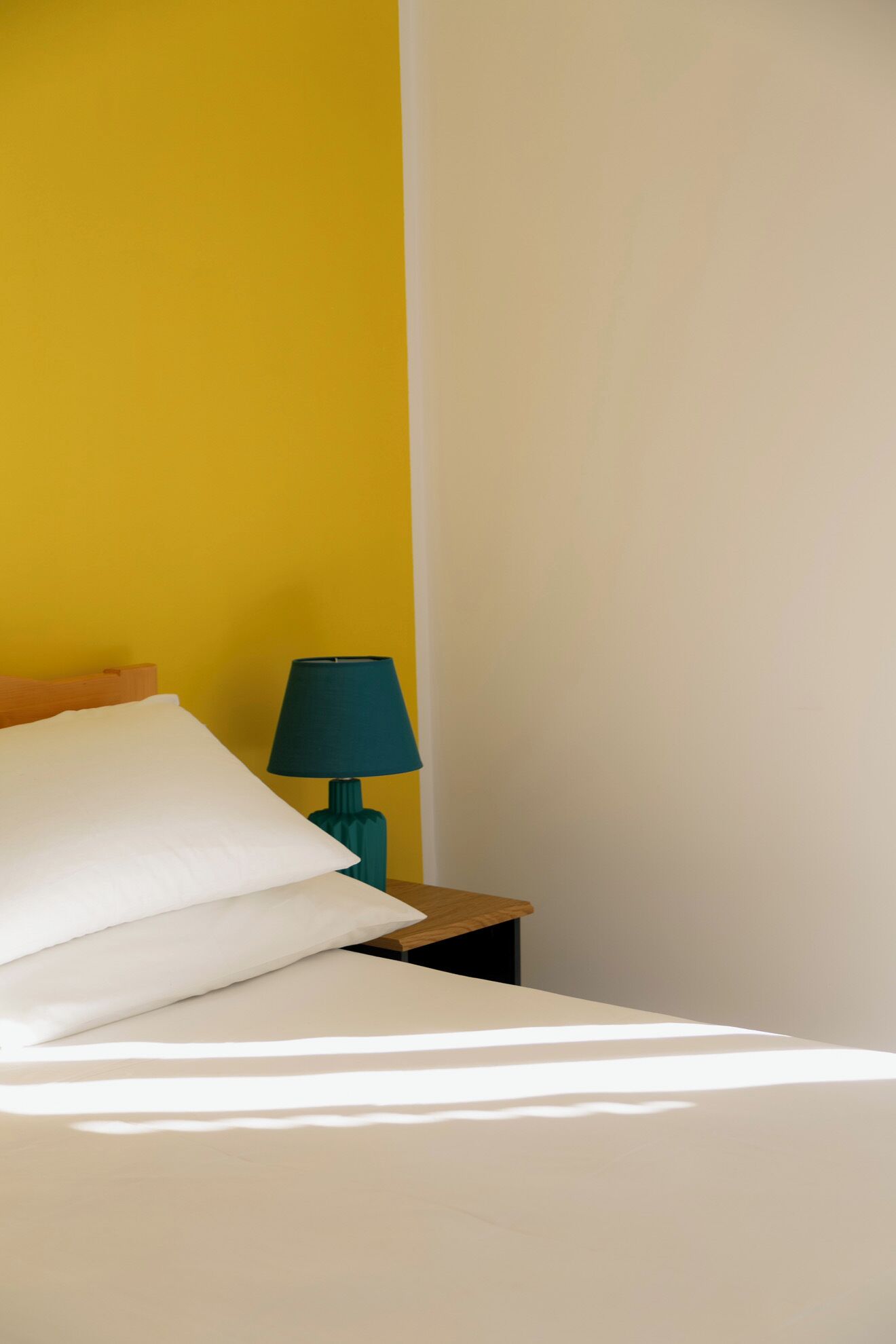 A bed with white linens, a teal lamp on a wooden nightstand, and a yellow wall.
