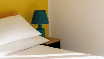A bed with white linens, a teal lamp on a wooden nightstand, and a yellow wall.