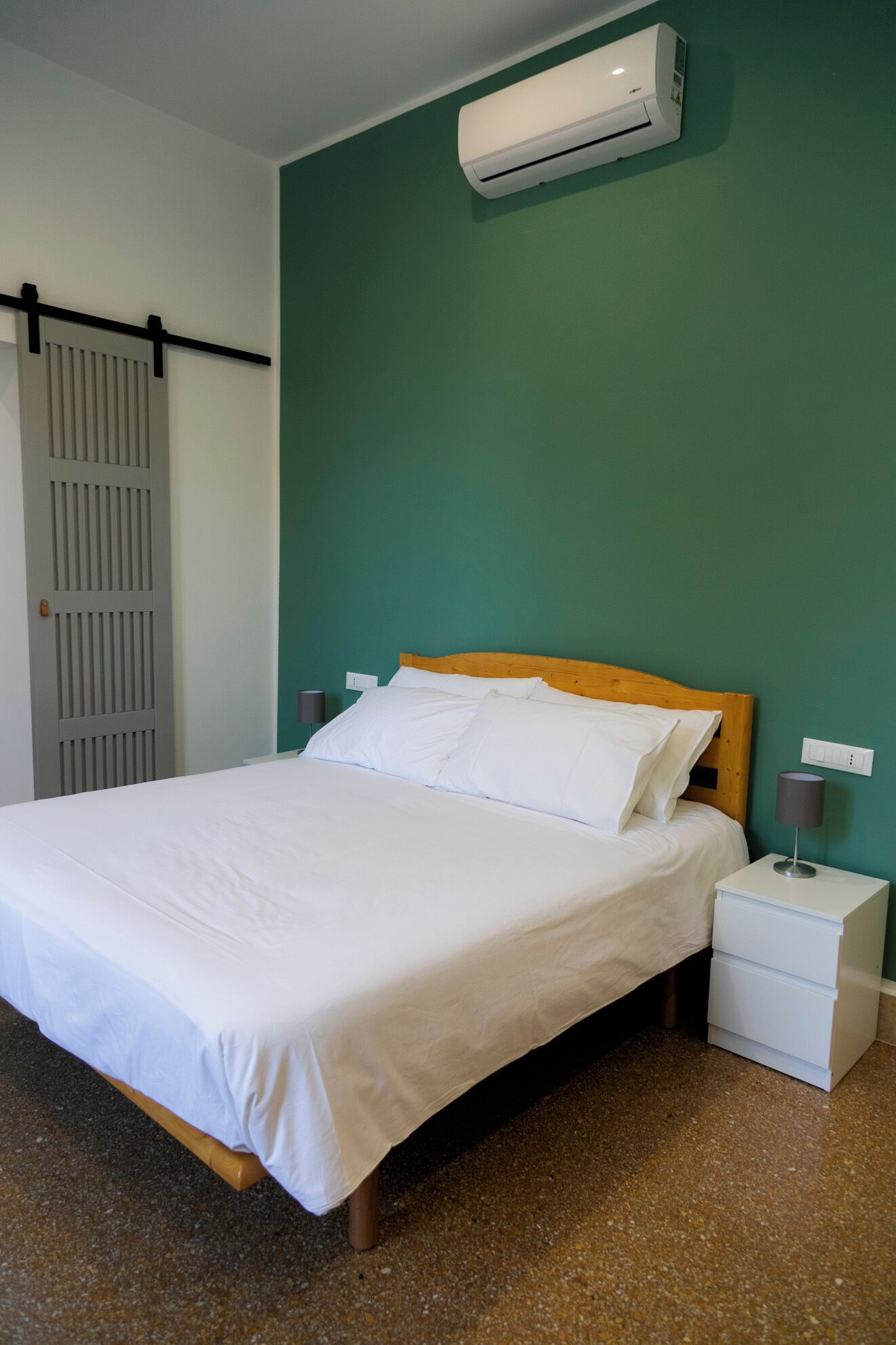 Un letto ben rifatto con lenzuola bianche, una testiera in legno, un comodino bianco con una lampada e un condizionatore d'aria fissato al muro.