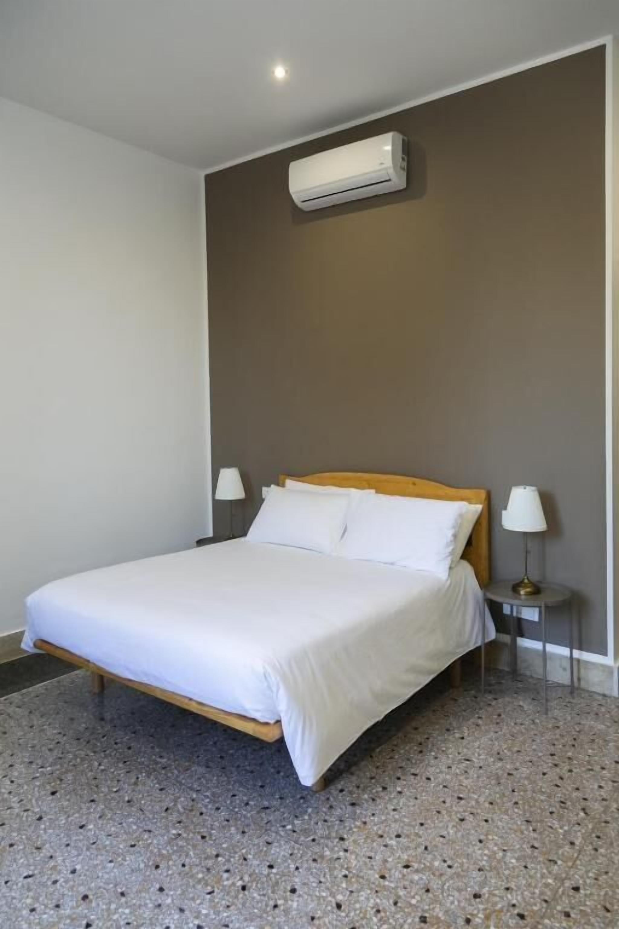 Quarto com cama, duas mesas de cabeceira, aparelho de ar condicionado e luminária na parede.