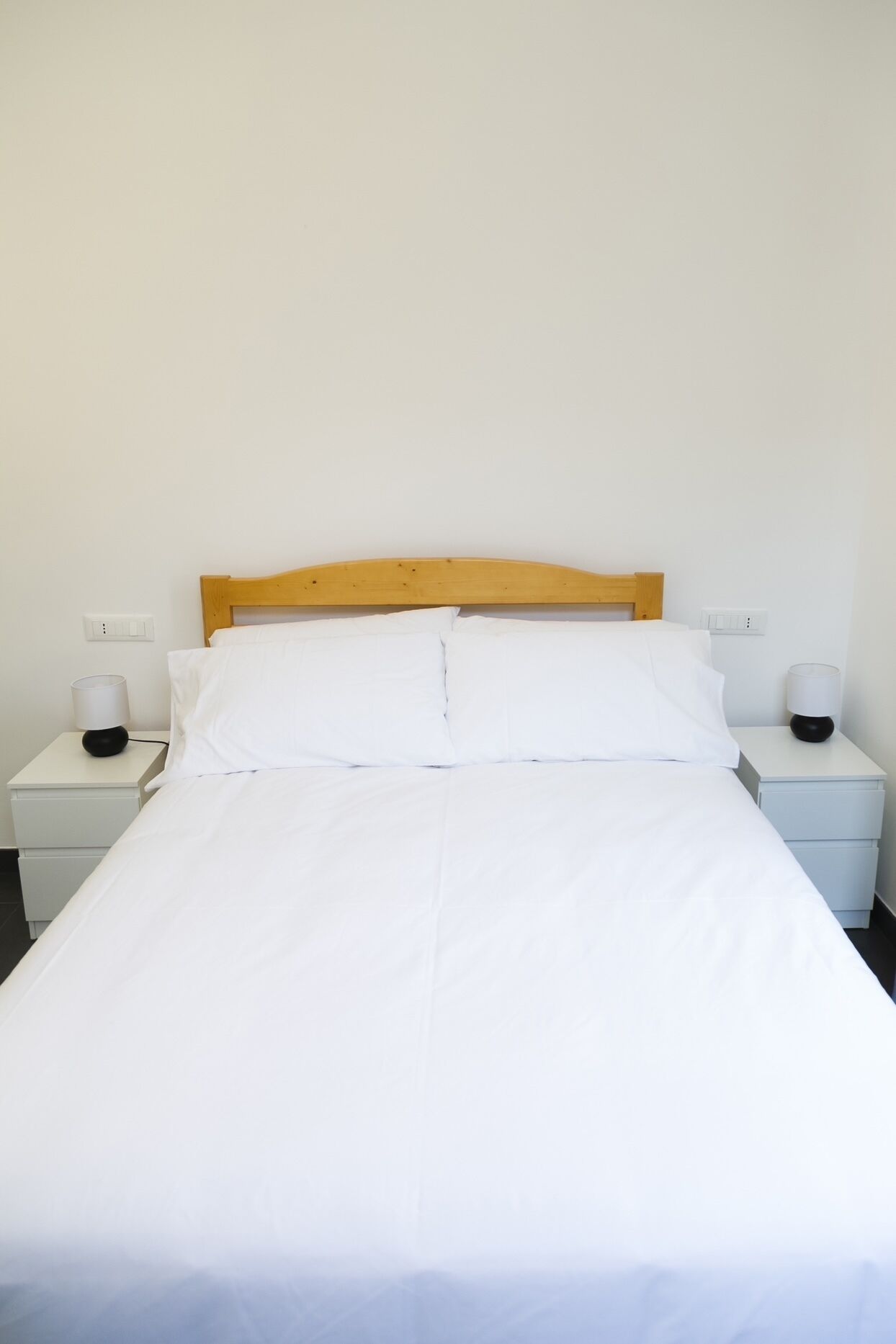 Cama bem arrumada com lençóis brancos, cabeceira de madeira e duas mesas de cabeceira com abajures.