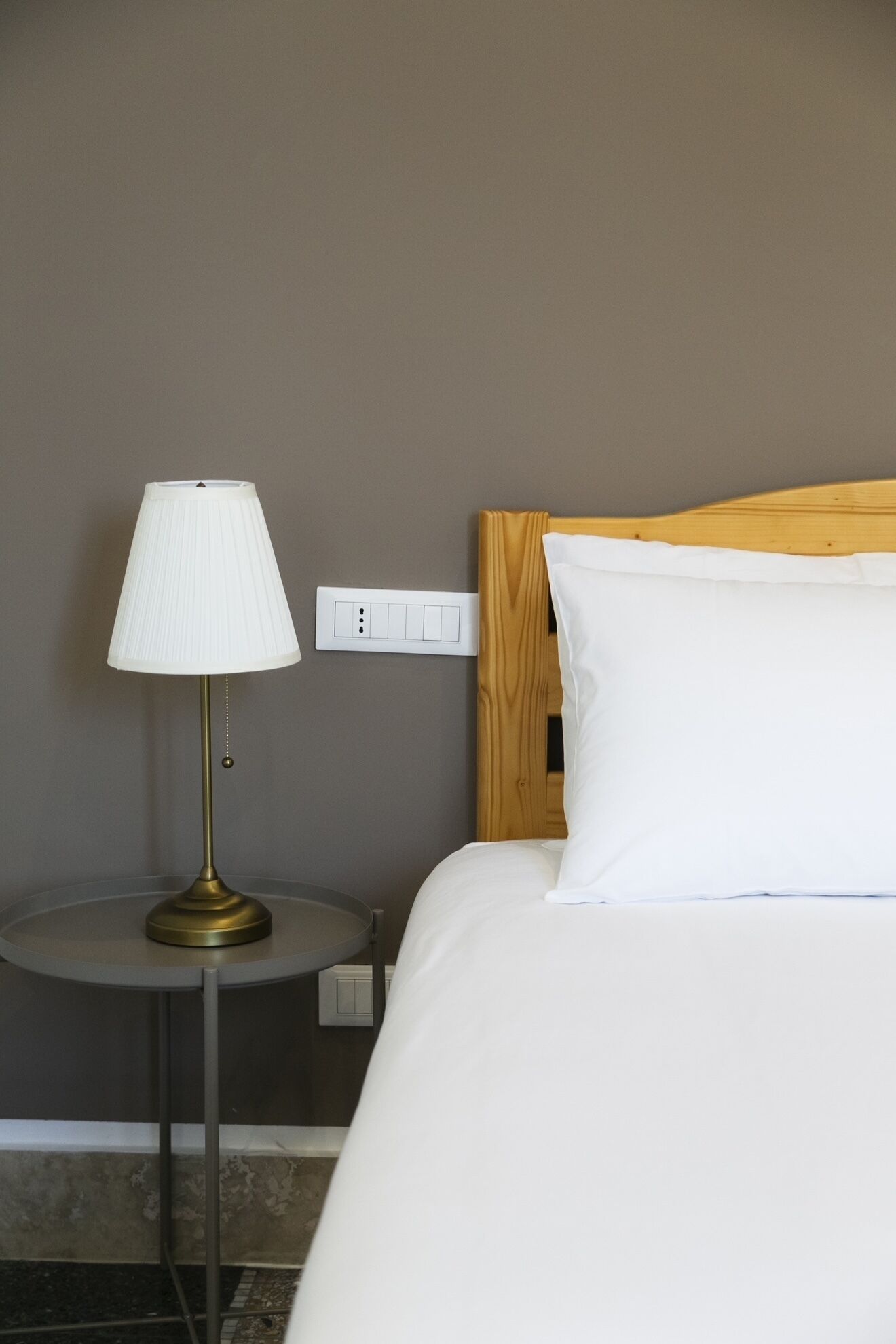 Hotellihuone, jossa on sänky, yöpöytälamppu ja pistorasia seinässä.