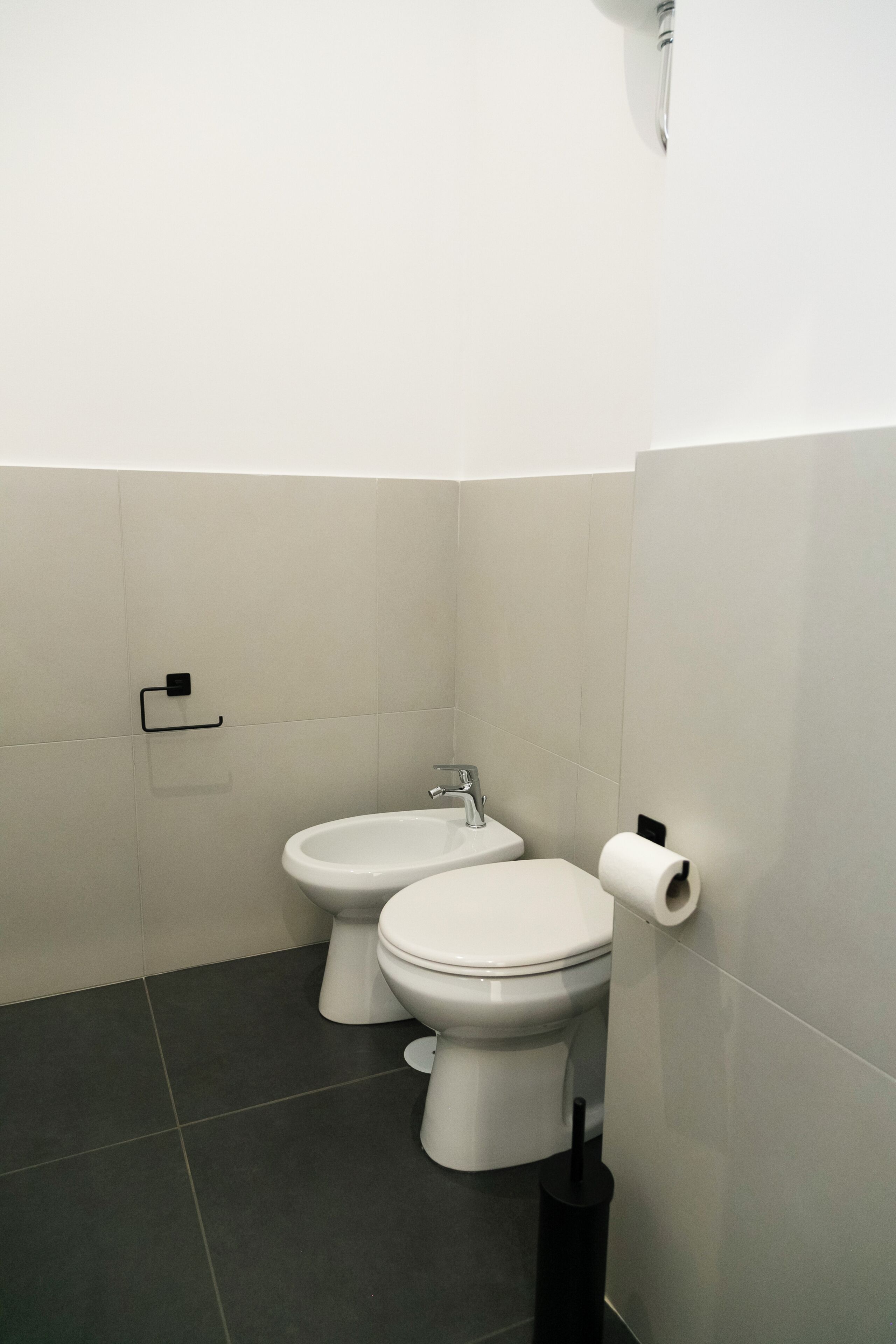 Un baño pequeño y moderno con dos inodoros, un lavamanos y un dispensador de papel higiénico.