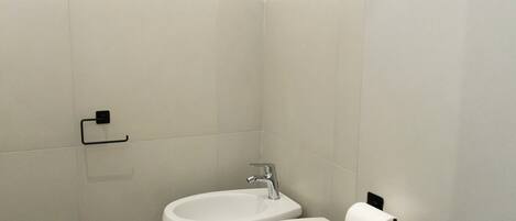 Un baño pequeño y moderno con dos inodoros, un lavamanos y un dispensador de papel higiénico.