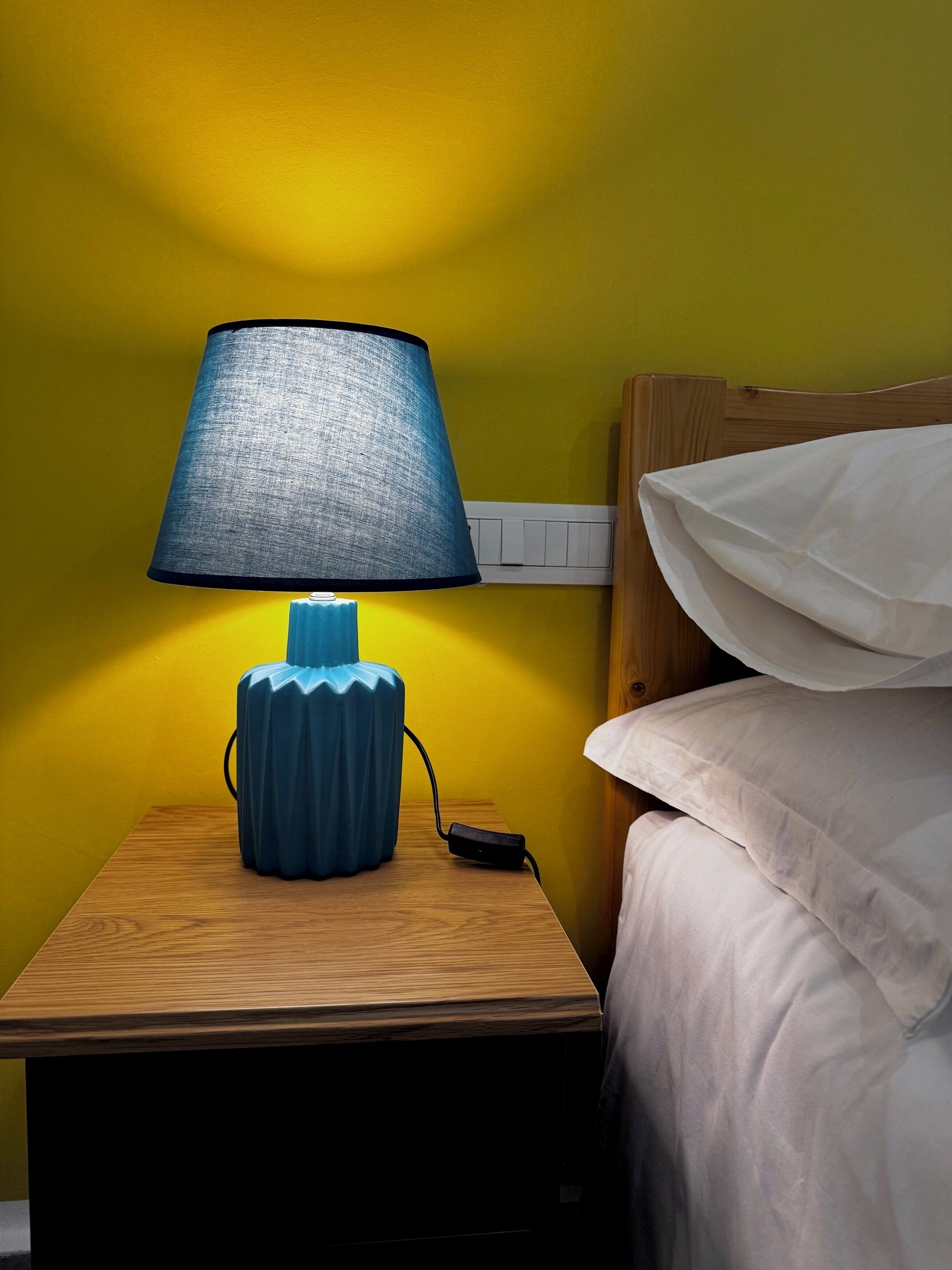 Una mesita de noche con una lámpara azul, una regleta y un almohadón blanco sobre un mueble de madera frente a una pared amarilla.