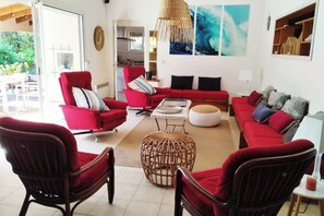 Un salon avec des meubles rouges, un pouf en osier et une grande fenêtre.