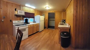 Una cocina compacta con paredes de madera, una estufa, un microondas, un refrigerador, un lavamanos y un cubo de basura.