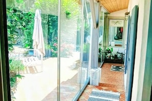 Une porte vitrée donne sur un patio dont le sol est recouvert de carreaux, où se trouve un parasol blanc et un chemin pavé en briques.