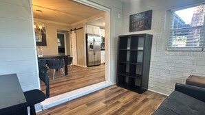 Een moderne woonkamer met een houten vloer, een donkergrijze bank, een eethoek met een tafel en stoelen, en een keuken met witte kastjes en een roestvrijstalen koelkast.
