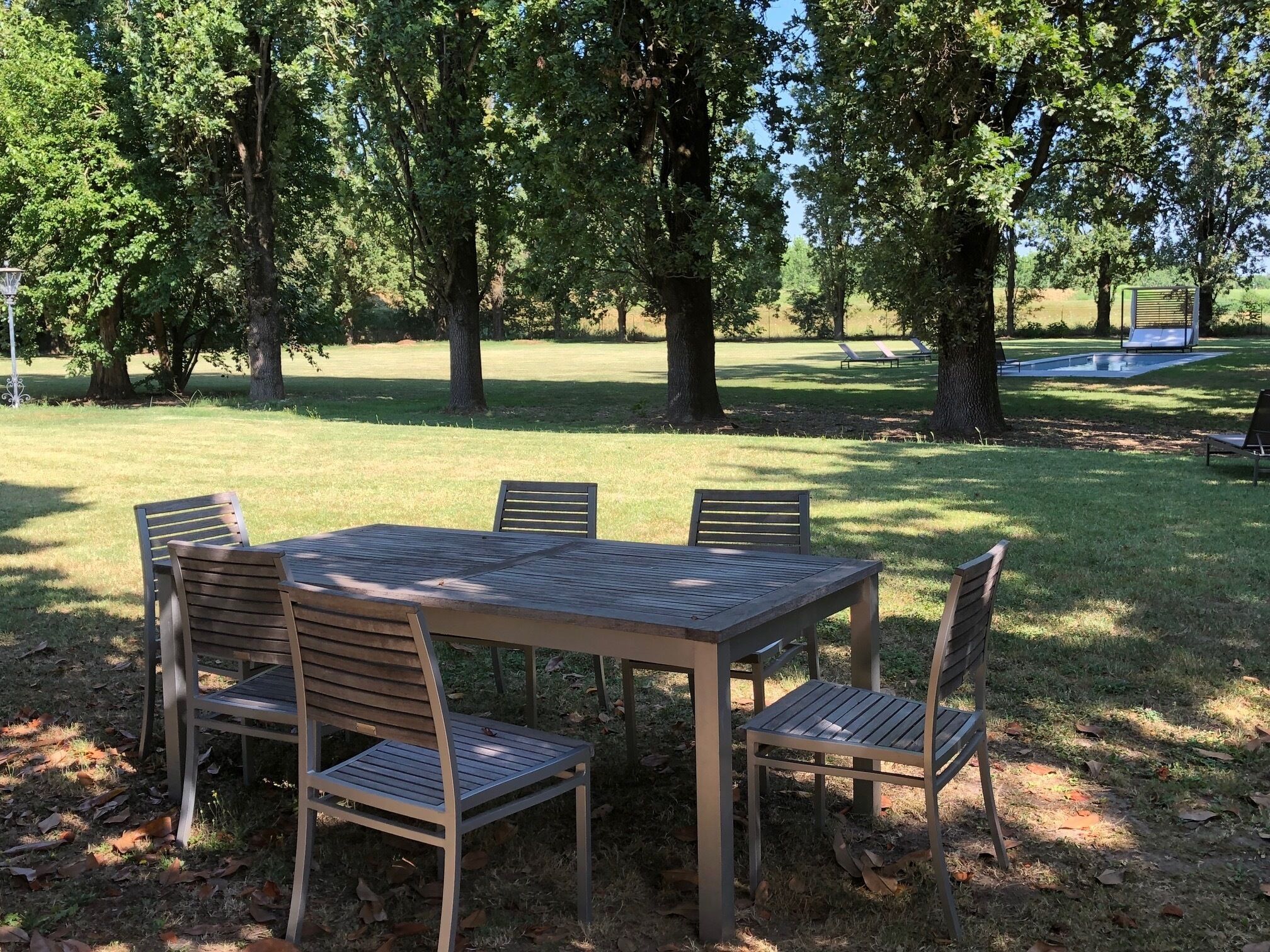 Una mesa de madera con cuatro sillas en un parque con árboles y una piscina al fondo.