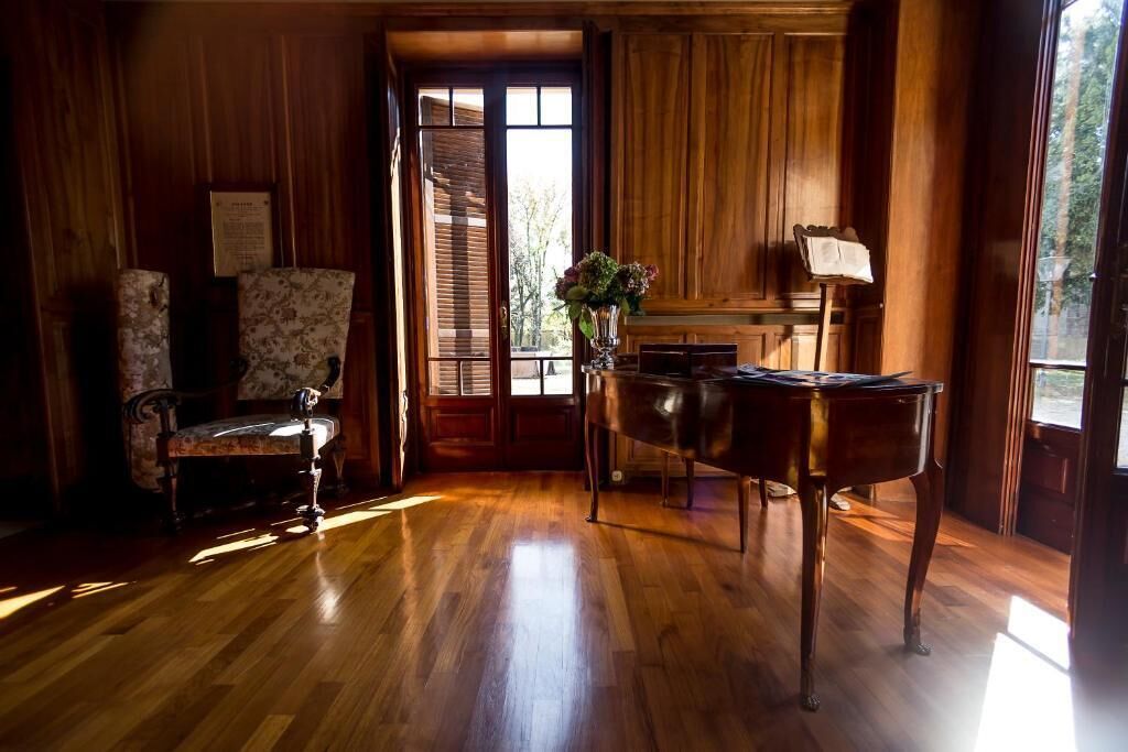 A room with wooden flooring, a piano, a chair, and a window with blinds.