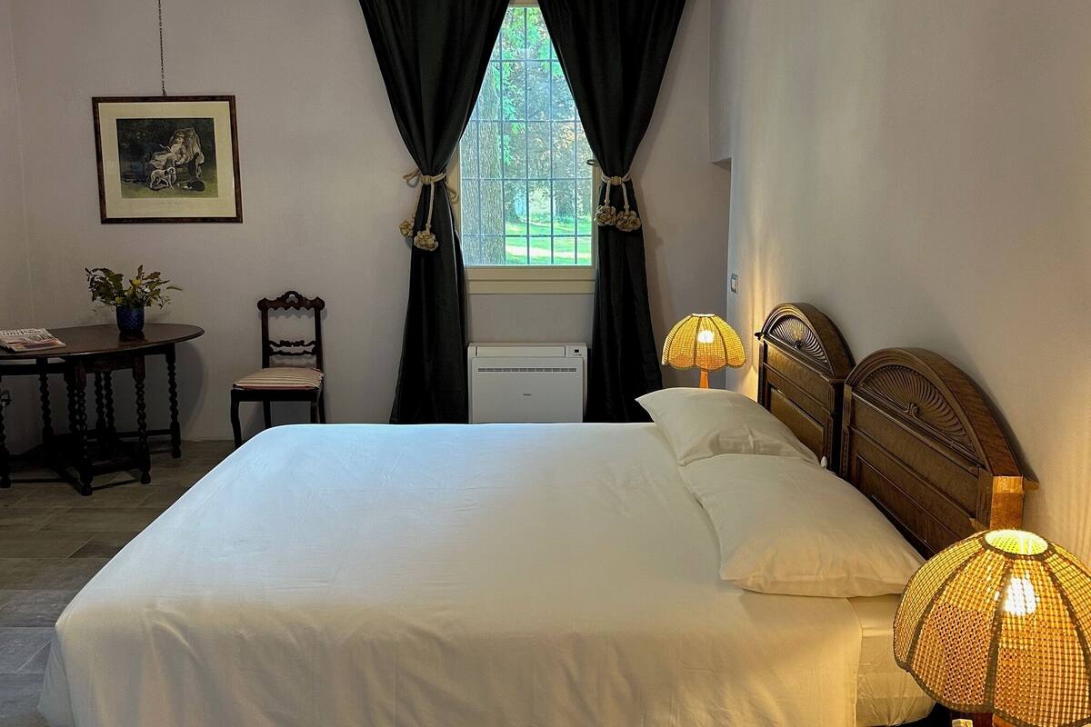 A bedroom with a bed, two bedside lamps, a window with curtains, a chair, a small table with a plant, and a framed picture on the wall.