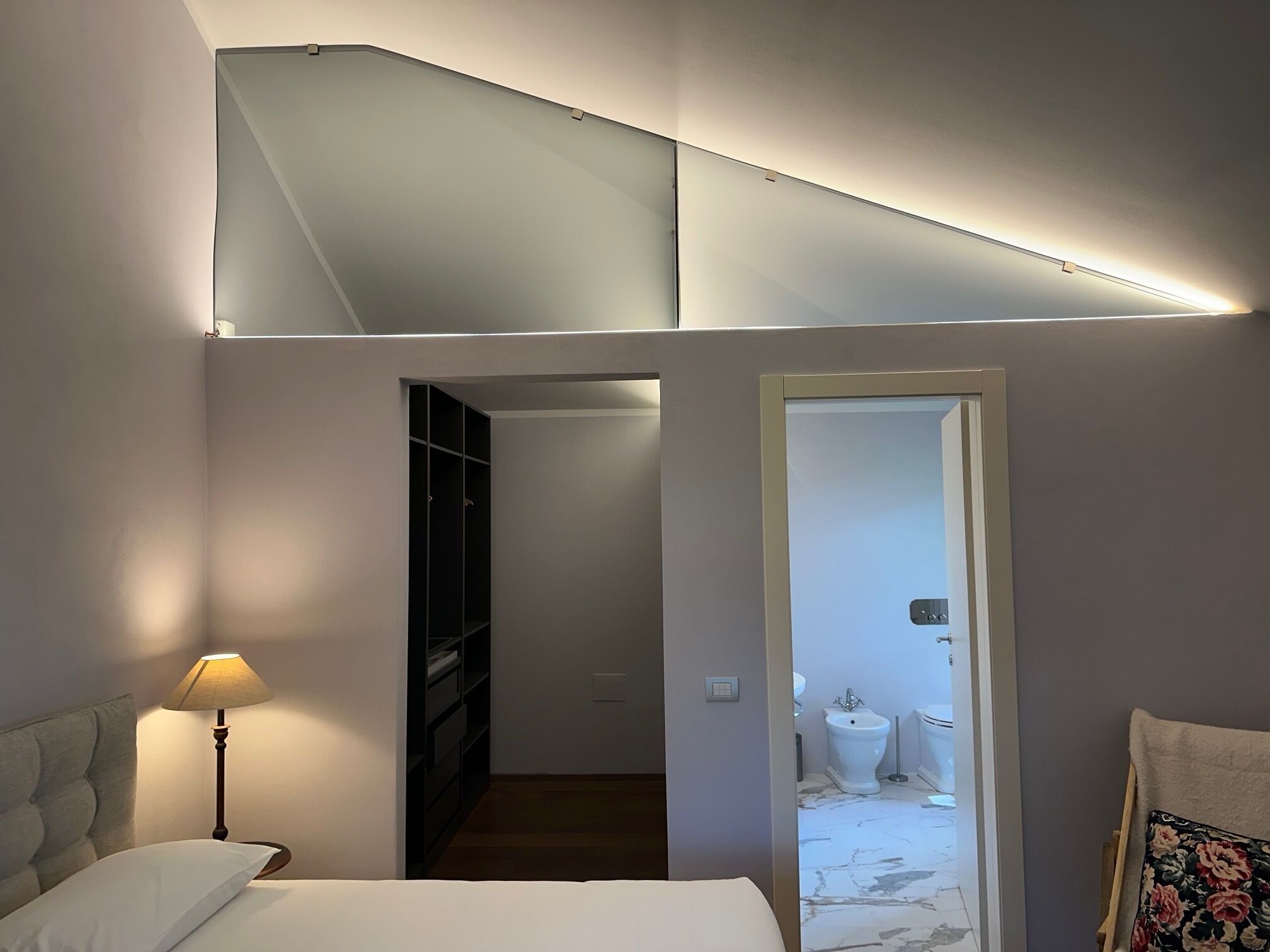 Un dormitorio moderno con cama, mesita de noche, lámpara, armario y un baño visible a través de una puerta abierta.