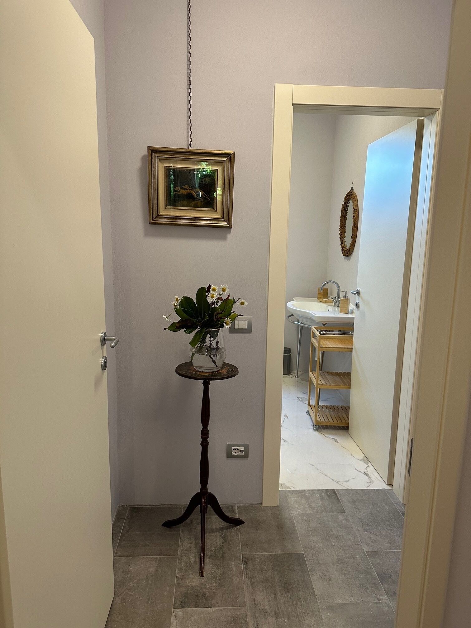Un couloir avec une porte, une petite table sur laquelle est posé un vase de fleurs, un tableau encadré au mur, et un aperçu d’une salle de bain avec un lavabo et des étagères.